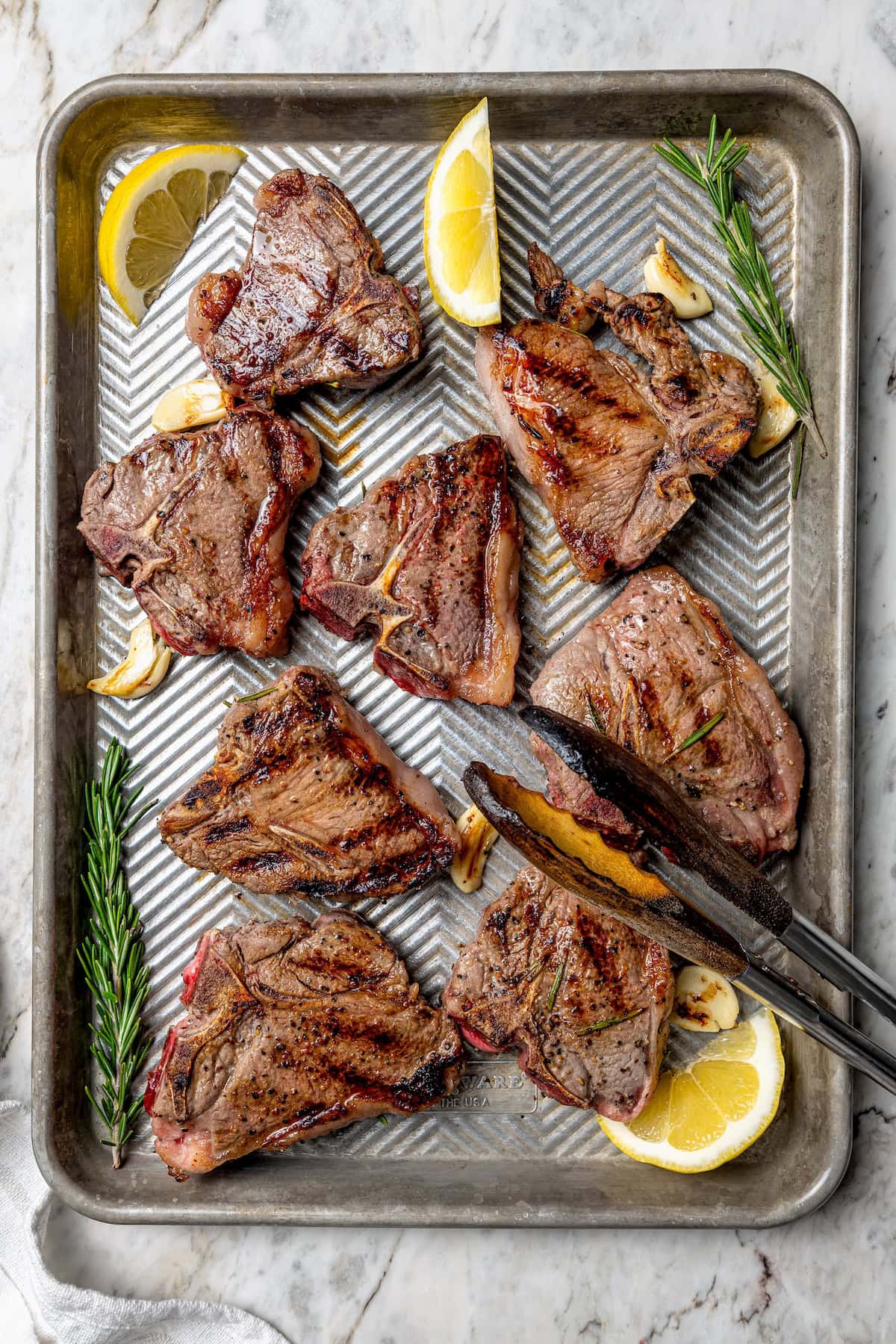 Overhead view of grilled lamb chops on sheet pan with fresh rosemary, lemon wedges, and garlic cloves