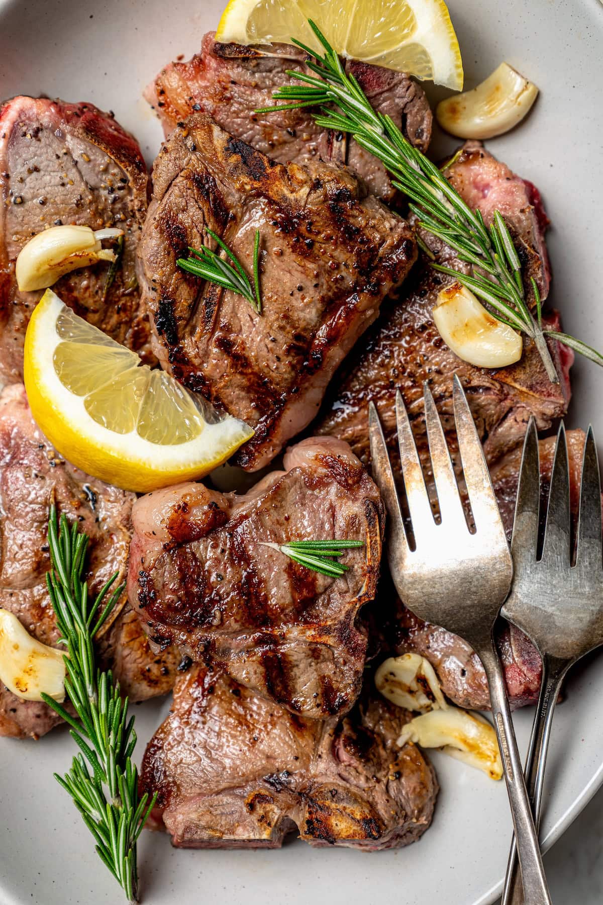 Overhead view of grilled lamb chops on platter with fresh rosemary, lemon wedges, and garlic cloves