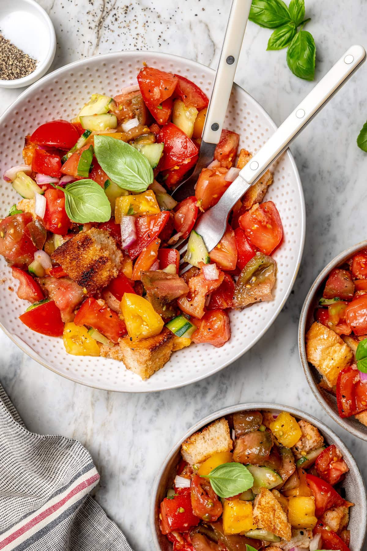 Overhead view of panzanella salad in serving bowl with fork and spoon and two smaller bowls