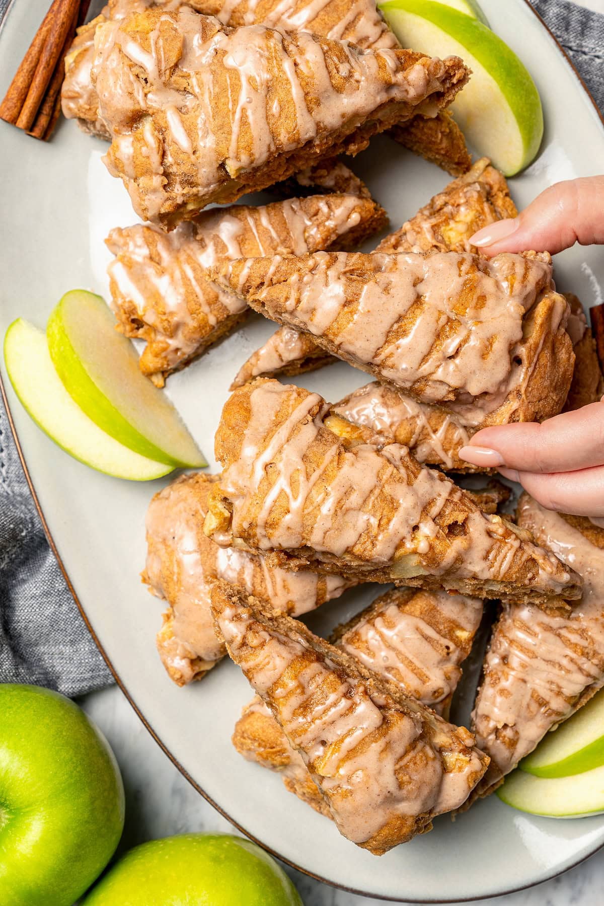 Buttermilk gives these spiced apple scones a moist, tender crumb, while applesauce replaces much of the butter to lighten them up! Hand grabbing glazed apple scone from platter