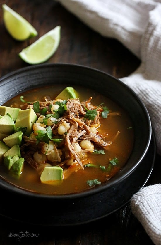 Bowl of pozole rojo with avocado and cilantro for garnish and lime wedges in background