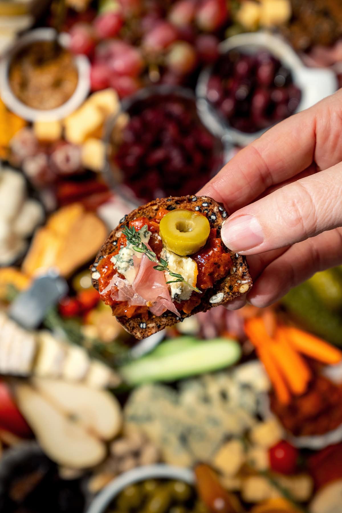 Hand holding cracker topped with cheese, meat, and olive.
